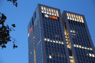Two tall office buildings with rectangular windows are lit up against a clear evening sky. The UBS logo is prominently displayed on the upper levels of both buildings. Some tree branches are visible on the left side.