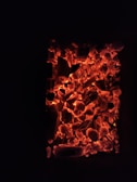 A bed of glowing embers with predominantly dark charcoal surrounded by bright red and orange hues, suggesting intense heat and burning. The image conveys a sense of warmth and energy.