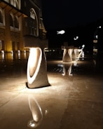 Illuminated stone structures stand in a row on a glossy, reflective surface. The lighting casts a warm glow on the structures and the surrounding area, enhancing the elegance of an adjacent building. The facade of the building is ornate, with archways and decorative elements. The overall atmosphere is serene and majestic, with additional illuminated features in the background.
