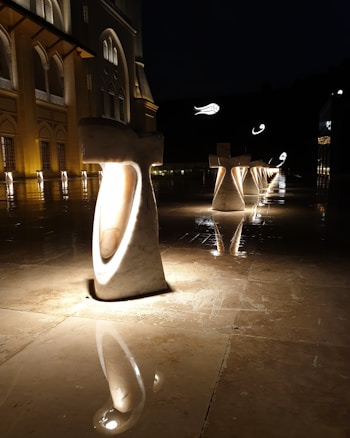 Illuminated stone structures stand in a row on a glossy, reflective surface. The lighting casts a warm glow on the structures and the surrounding area, enhancing the elegance of an adjacent building. The facade of the building is ornate, with archways and decorative elements. The overall atmosphere is serene and majestic, with additional illuminated features in the background.