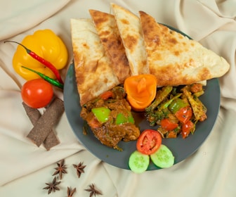 A dark plate filled with a colorful curry dish featuring assorted vegetables such as green and red peppers, broccoli, and a mix of spices, accompanied by several pieces of flatbread and garnished with sliced tomatoes and cucumber. Adjacent to the plate, there is a display of whole spices and fresh vegetables, including a yellow bell pepper, red and green chilies, a tomato, cinnamon sticks, and star anise, all placed on a cream-colored fabric background.