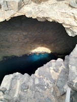 A natural rock formation with a large cavernous opening, leading to clear blue water at the bottom. The surrounding rock surfaces have a rugged texture, with light and shadow creating depth and contrast.