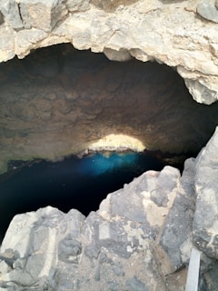 A natural rock formation with a large cavernous opening, leading to clear blue water at the bottom. The surrounding rock surfaces have a rugged texture, with light and shadow creating depth and contrast.
