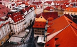 Aerial view of a historic cityscape featuring numerous ornate buildings with distinct red-tiled roofs, narrow streets, and a few people walking. The architecture presents a picturesque and traditional European style, with various shapes and interesting designs.