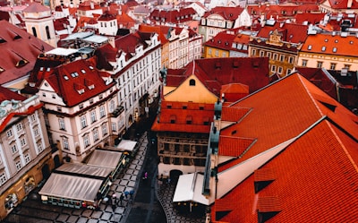 Aerial view of a historic cityscape featuring numerous ornate buildings with distinct red-tiled roofs, narrow streets, and a few people walking. The architecture presents a picturesque and traditional European style, with various shapes and interesting designs.