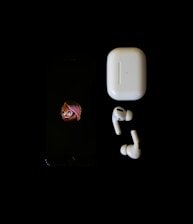 A smartphone displaying an emoji character is placed next to a pair of wireless earbuds and their charging case against a black background.