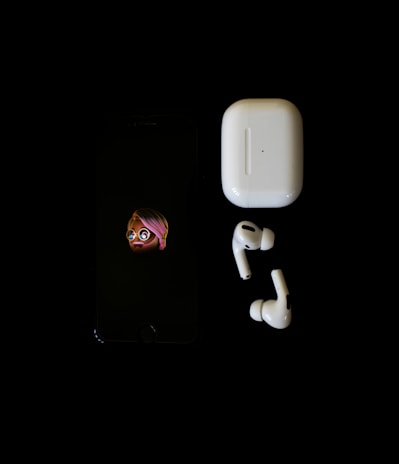 A smartphone displaying an emoji character is placed next to a pair of wireless earbuds and their charging case against a black background.