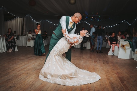 A couple is gracefully dancing together on a wooden dance floor, with the man in a green suit and the woman in a white lace bridal gown. They are surrounded by seated guests and ambient string lights, creating a festive atmosphere.