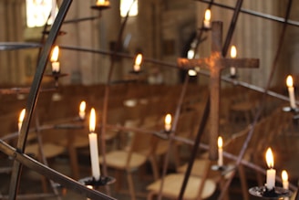 A series of lit candles is arranged on a metal holder in the foreground, with a blurred background of wooden chairs and what appears to be a church or cathedral interior. The soft lighting creates a serene and contemplative atmosphere.