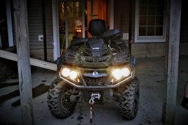 A rugged all-terrain vehicle (ATV) with bright headlights is parked in a covered area outside a house. The ATV appears muddy, indicating recent off-road use. The structure has wooden beams and a wooden ramp is visible on the left side.