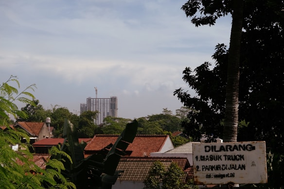 A view of a residential area with red-tiled roofs and lush greenery in the foreground. In the background, a tall building under construction is visible against a partly cloudy sky. There is a sign in the lower right corner with some text.