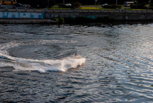 A person on a jet ski creates a large spray as they maneuver on a wide body of water. The water is slightly rippled with reflections of the sky and surrounding environment. In the background, there is a long wall with graffiti, a grassy area, and a few parked vehicles.
