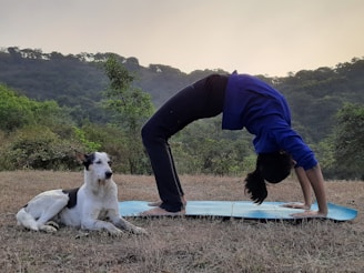 A person performs a yoga backbend on a blue mat outdoors, with a black and white dog sitting calmly nearby. The setting is a grassy area surrounded by lush green hills and trees under a calm sky.