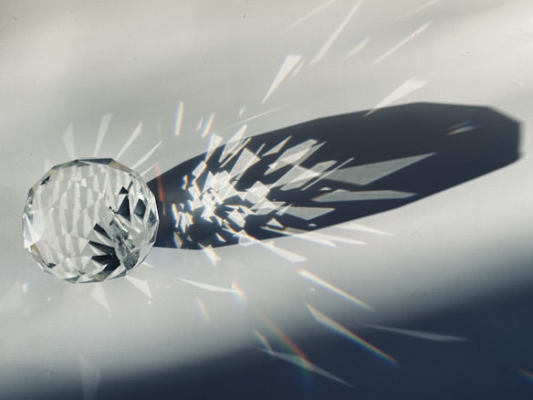 A multifaceted crystal ball lies on a surface under bright lighting, casting intricate shadows and refracted light patterns that resemble starbursts. The surface and lighting create a dramatic interplay of light and shadow.