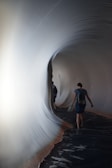 A person walks through a tunnel-like structure with smooth, curving surfaces that resemble fabric or stretched material. The interior is dimly lit, with a soft glow illuminating the contours of the tunnel, creating an immersive and surreal environment.
