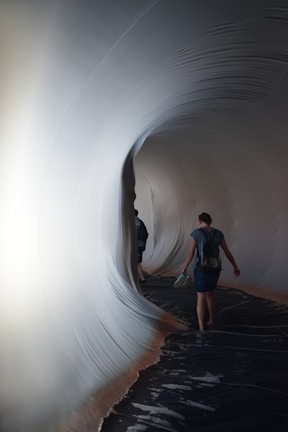 A person walks through a tunnel-like structure with smooth, curving surfaces that resemble fabric or stretched material. The interior is dimly lit, with a soft glow illuminating the contours of the tunnel, creating an immersive and surreal environment.