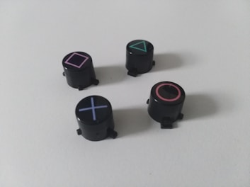 Four black cylindrical objects with distinct geometric symbols: a pink square, a green triangle, a red circle, and a blue cross. These symbols are associated with gaming console controllers.