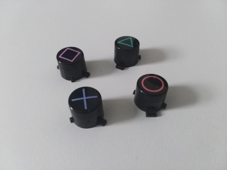 Four black cylindrical objects with distinct geometric symbols: a pink square, a green triangle, a red circle, and a blue cross. These symbols are associated with gaming console controllers.