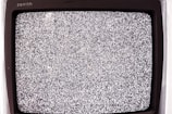 A close-up view of an old television screen displaying static noise. The screen is filled with a pattern of black and white dots and spots, typical of a signal loss or disturbance. The television set has a vintage design with a black frame.