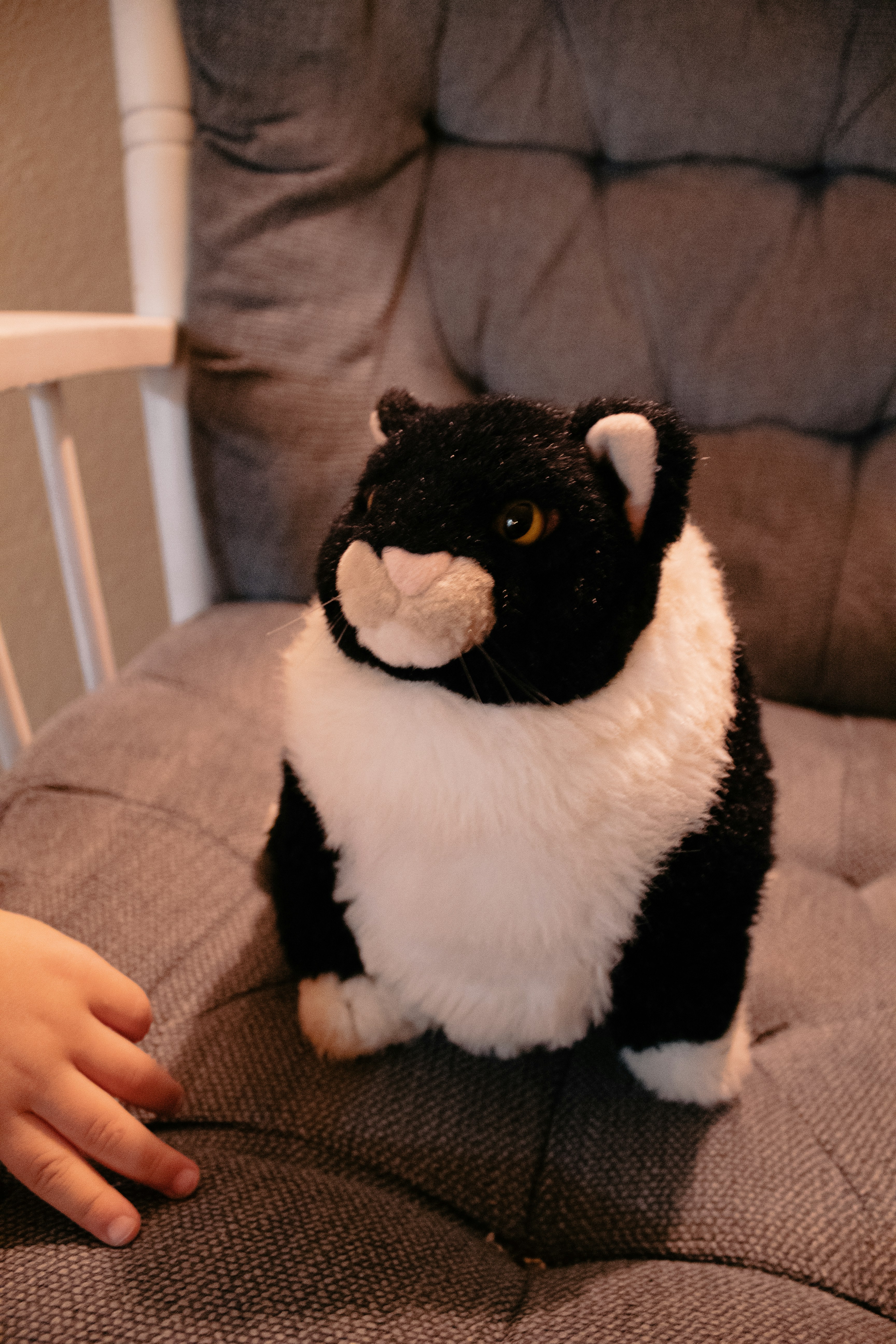 A plush toy resembling a black and white cat sits on a cushioned chair. A small hand reaches towards the toy, suggesting the presence of a child.