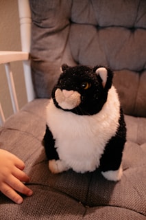 A plush toy resembling a black and white cat sits on a cushioned chair. A small hand reaches towards the toy, suggesting the presence of a child.