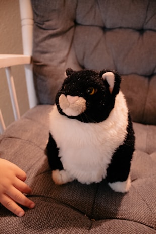 A plush toy resembling a black and white cat sits on a cushioned chair. A small hand reaches towards the toy, suggesting the presence of a child.