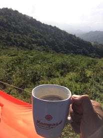 A person is holding a white cup with a logo against a backdrop of lush, green hills and a partly cloudy sky. The foreground features an orange fabric, possibly part of a tent or camping gear.