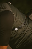 A close-up of a person wearing a dark, textured jacket or vest with a strap over the shoulder. The label 'RILLE' is visible on the fabric, and there's a part of a camera or equipment with a strap attached.