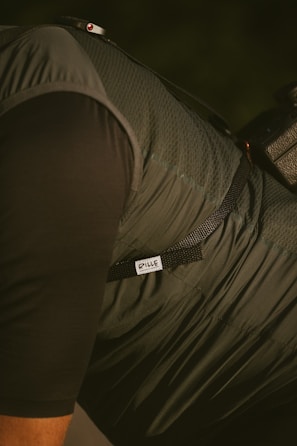 A close-up of a person wearing a dark, textured jacket or vest with a strap over the shoulder. The label 'RILLE' is visible on the fabric, and there's a part of a camera or equipment with a strap attached.