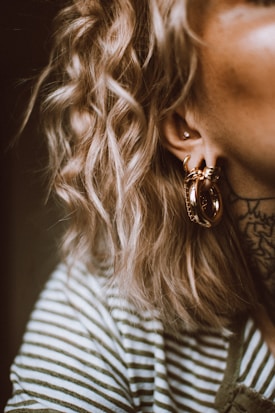 The image features a close-up of a person's ear adorned with large, gold hoop earrings. The person has wavy, blonde hair and wears a striped shirt. A tattoo is visible on the neck.