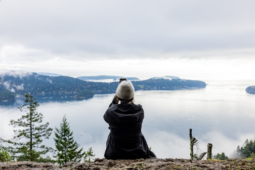 A person is sitting on a rocky ledge, photographing the scenic view of a serene body of water surrounded by forested islands. The sky is overcast, contributing to a peaceful and contemplative atmosphere.