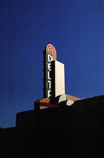 A retro-style theater marquee with the word 'Delta' prominently displayed in vertical letters on a large sign against a deep blue sky. The sign features red, green, and white colors with an art deco design.