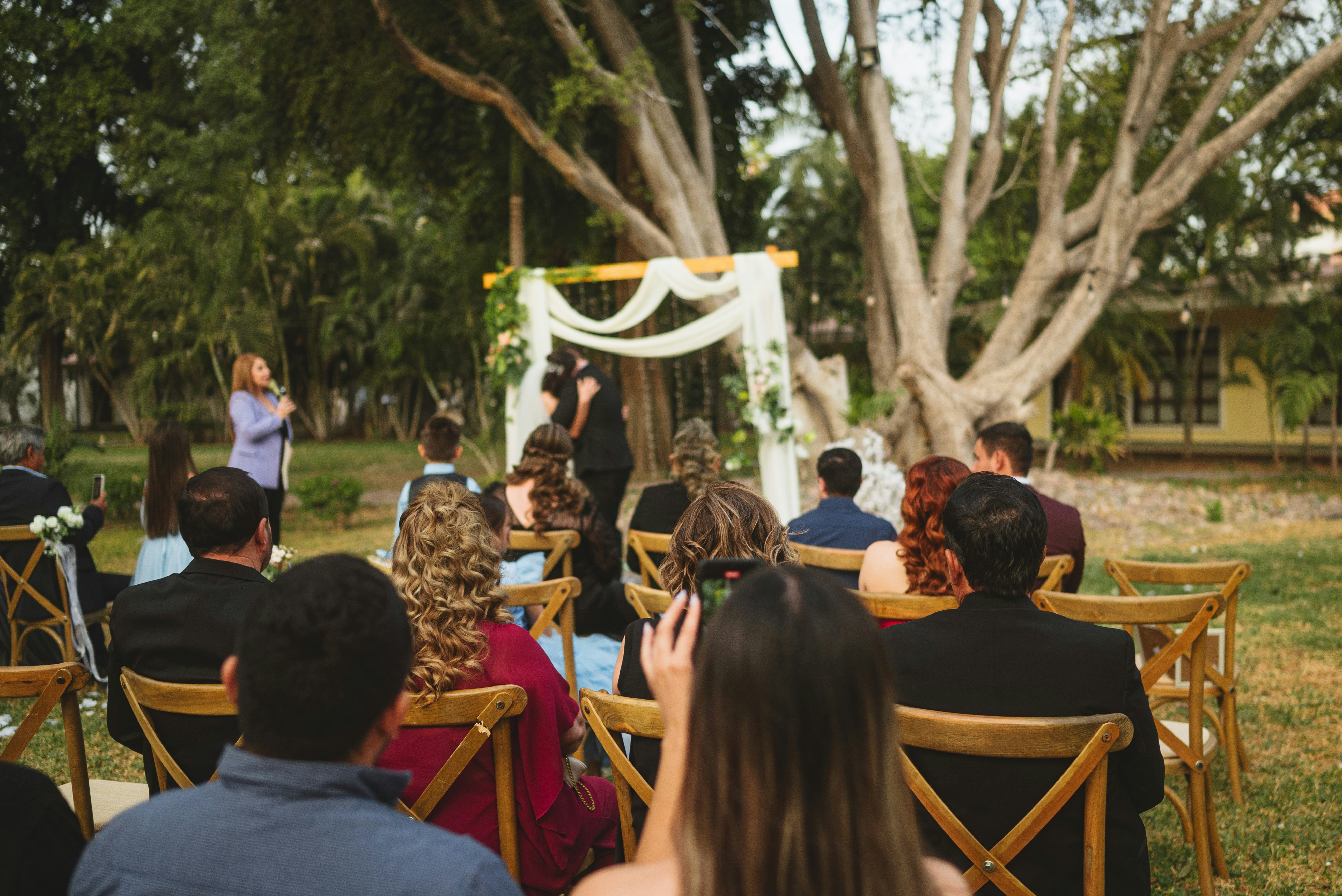 Convidados sentados em cadeiras de madeira assistindo a uma cerimônia de casamento em um jardim.
