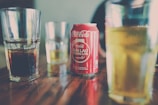 A red can of Coca-Cola with some Vietnamese text on it is placed on a wooden table surrounded by several glass tumblers containing different drinks.