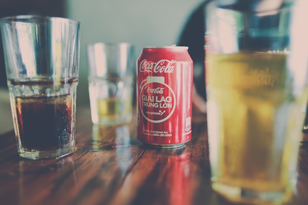 A red can of Coca-Cola with some Vietnamese text on it is placed on a wooden table surrounded by several glass tumblers containing different drinks.