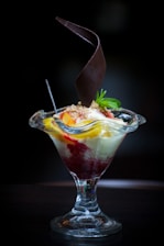 A dessert served in a tall glass dish with a combination of vanilla ice cream, fruit toppings, and a drizzle of red sauce. A decorative chocolate piece spirals upwards from the center, and a mint sprig garnishes the top.