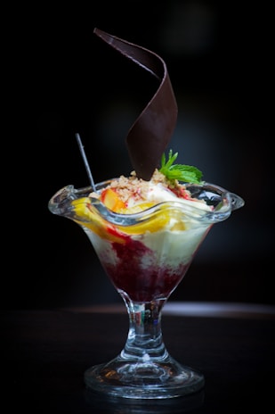 A dessert served in a tall glass dish with a combination of vanilla ice cream, fruit toppings, and a drizzle of red sauce. A decorative chocolate piece spirals upwards from the center, and a mint sprig garnishes the top.