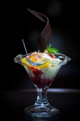 A dessert served in a tall glass dish with a combination of vanilla ice cream, fruit toppings, and a drizzle of red sauce. A decorative chocolate piece spirals upwards from the center, and a mint sprig garnishes the top.