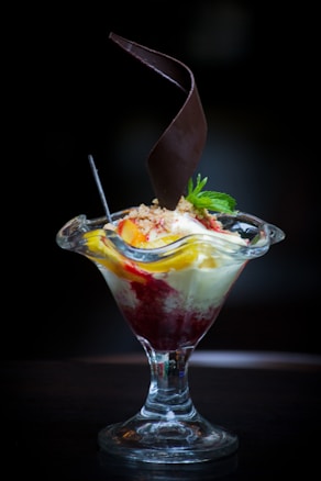 A dessert served in a tall glass dish with a combination of vanilla ice cream, fruit toppings, and a drizzle of red sauce. A decorative chocolate piece spirals upwards from the center, and a mint sprig garnishes the top.