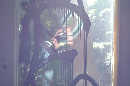 A glass window features dripping black paint in abstract shapes. Behind the glass, a person holding a camera is partially visible due to the reflective surface. Sunlight streams in, casting reflections and creating a hazy, artistic atmosphere.