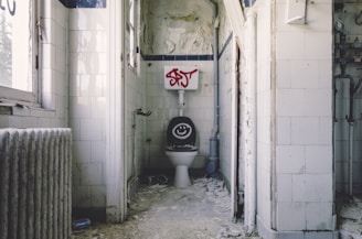 A small, neglected bathroom with peeling paint and graffiti on the walls. The toilet seat is open, revealing a smiley face design on the lid. The floor is covered with debris and dirt. The walls are tiled in white, and there are visible pipes along with an old radiator near the window.