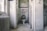 A small, neglected bathroom with peeling paint and graffiti on the walls. The toilet seat is open, revealing a smiley face design on the lid. The floor is covered with debris and dirt. The walls are tiled in white, and there are visible pipes along with an old radiator near the window.