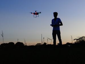 A silhouette of a person operating a drone in an open field at dusk. The sky is a gradient of blue to orange, with trees and poles in the background. The drone is illuminated with red and green lights.