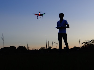 A silhouette of a person operating a drone in an open field at dusk. The sky is a gradient of blue to orange, with trees and poles in the background. The drone is illuminated with red and green lights.