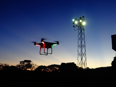 A drone with red and green lights is silhouetted against a darkening sky, with an industrial light tower illuminated in the background. Trees are visible at the base of the image, casting shadows as the sun sets.
