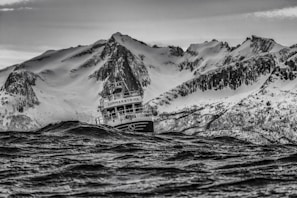 A fishing boat navigates through choppy waters with a backdrop of snow-covered mountains. The scene captures a moment of rugged natural beauty, with the boat appearing to struggle against the force of the waves.