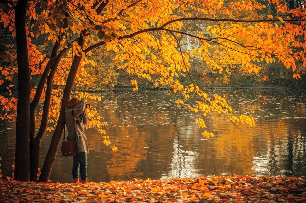 woman in brown coat standing near orange leafed tree and body of water during daytime