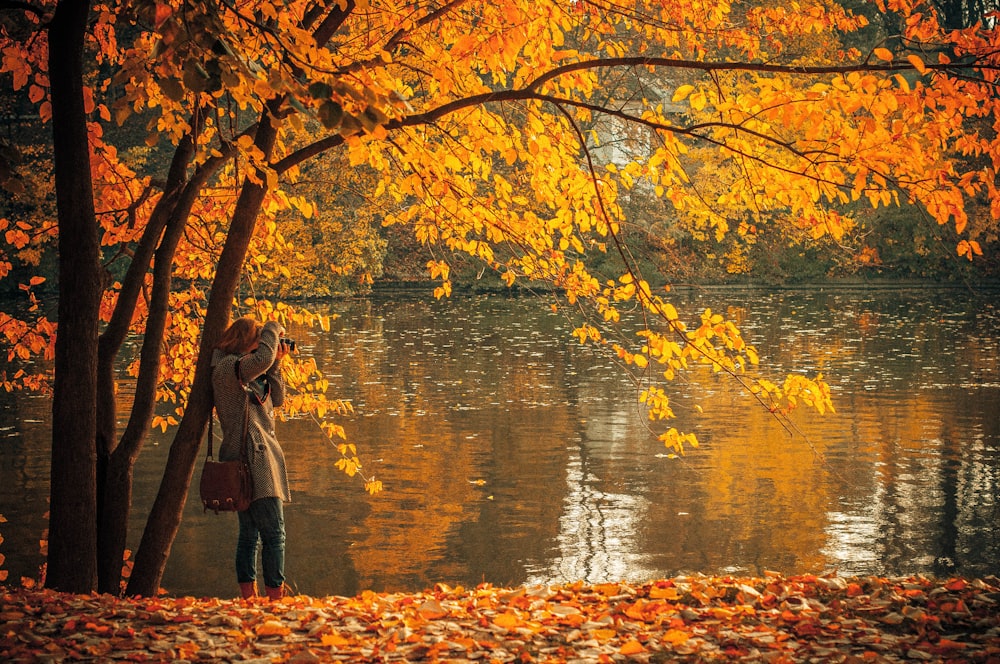 日中、オレンジ色の葉の木と水域のそばに立つ茶色のコートを着た女性