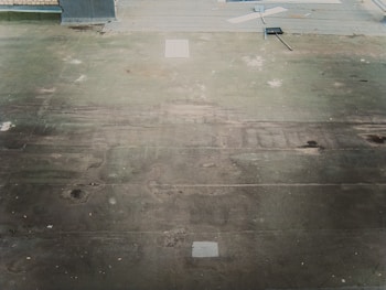 A rooftop surface with a worn and weathered appearance, featuring dirt and debris scattered across. A broom and a dustpan with a long handle lie on the roof, suggesting recent or ongoing cleaning activity. The surface has patches of discoloration, and there are some papers or objects near the top edge.