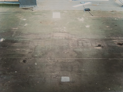 A rooftop surface with a worn and weathered appearance, featuring dirt and debris scattered across. A broom and a dustpan with a long handle lie on the roof, suggesting recent or ongoing cleaning activity. The surface has patches of discoloration, and there are some papers or objects near the top edge.